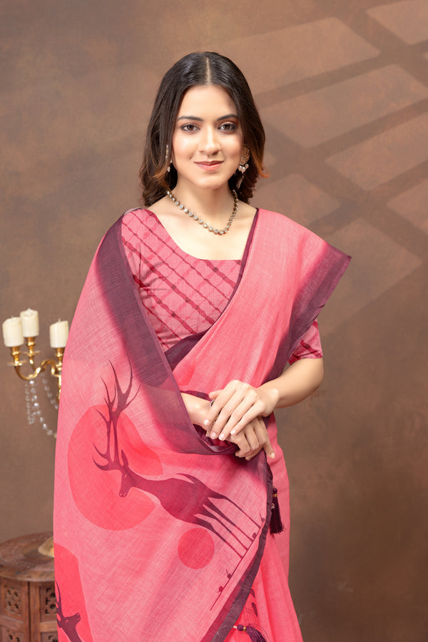 A woman wearing a pink and red printed saree with a matching blouse, standing against a brown textured studio backdrop with a candelabra partially visible on the left.